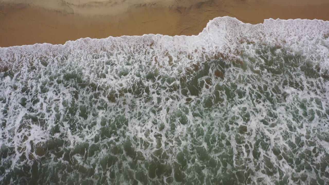 Aerial Los Cabos shoreline frame with white bubbled foam stretching horizontally across the beach edge and no visible people.