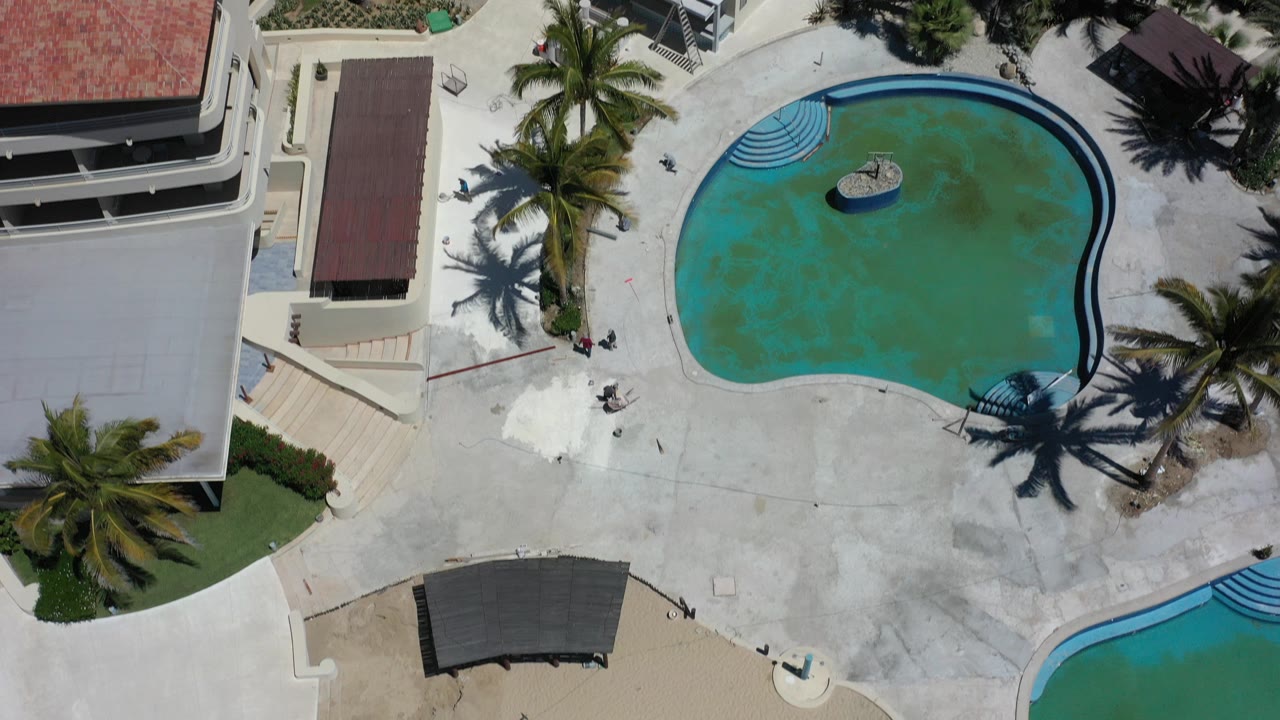 Overhead view in Los Cabos, Mexico, showing a curved turquoise resort pool, pale deck, palm trees, red tile roofs, and no visible people at the opening moment.