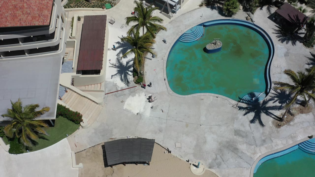 Aerial overhead frame in Los Cabos, Mexico, with the kidney-shaped pool slightly right of center, palm shadows across the deck, resort buildings around it, and no visible people.