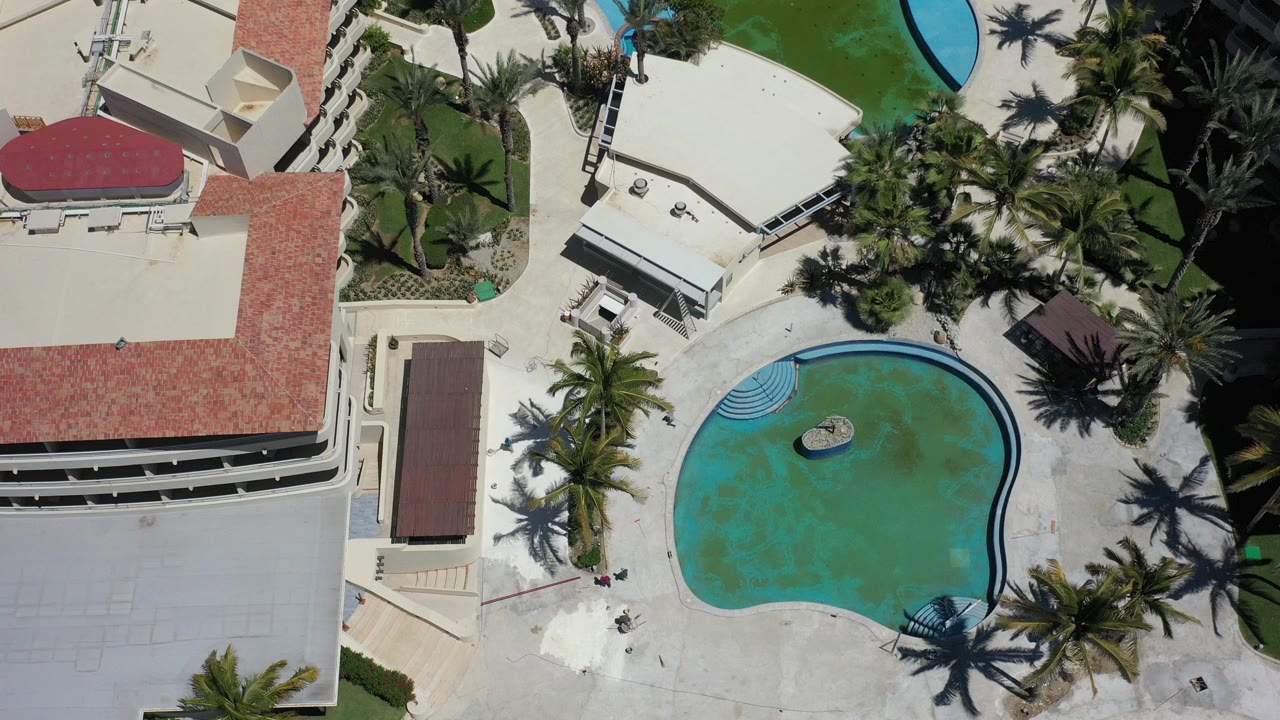 Overhead view in Los Cabos, Mexico, showing a turquoise curved pool beside cream hotel buildings with red tile roofs, palm landscaping, and no visible people.