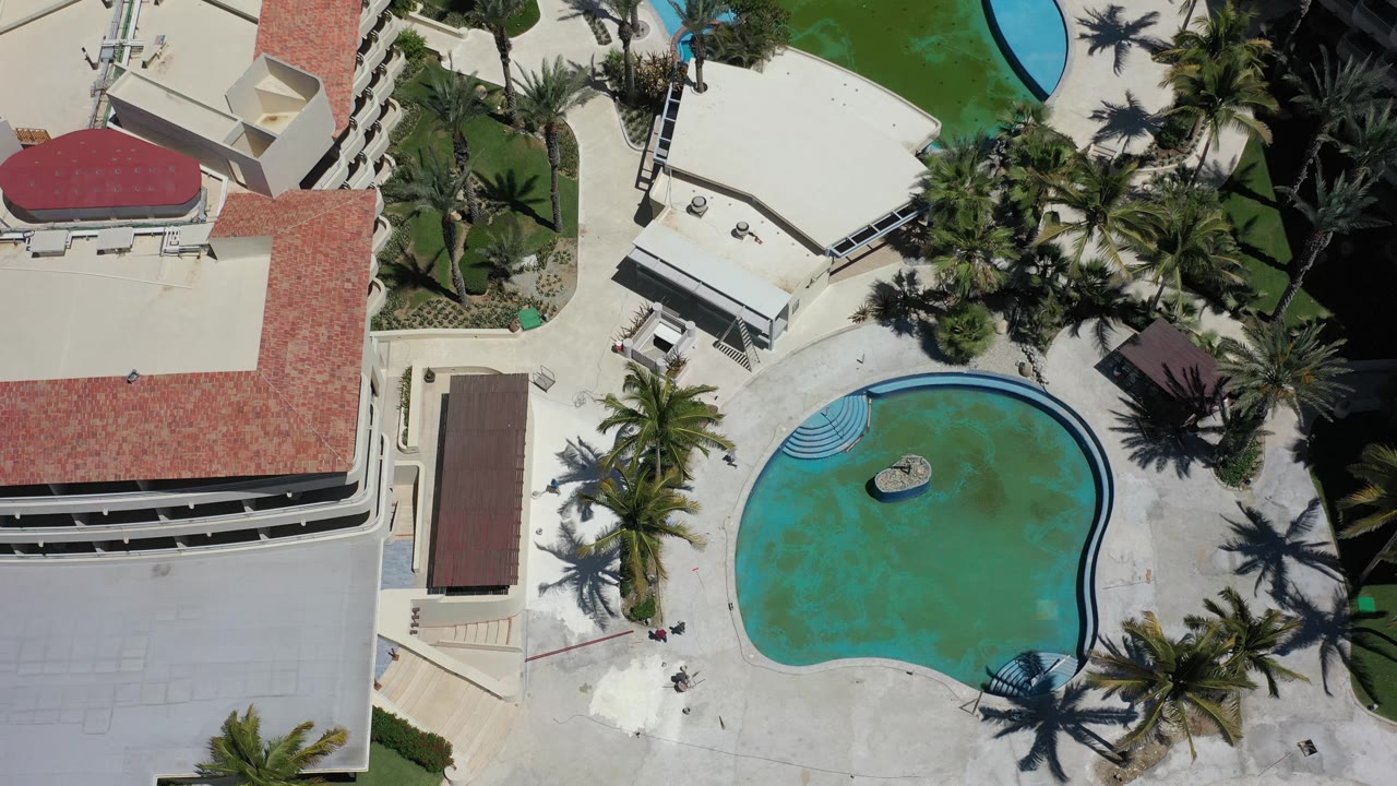 Aerial frame over a Los Cabos resort pool, with the oval pool centered lower right, red-roof hotel structures to the left, palm shadows on the deck, and no visible people.