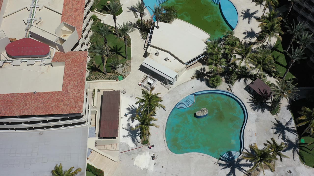 Top-down resort pool beside hotel buildings and palm landscaping.