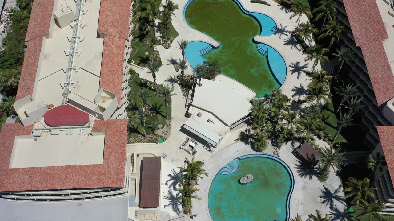 Overhead view in Los Cabos, Mexico showing curved turquoise resort pools, pale decks, red-tile roofs, cream buildings, palm landscaping, and no visible people.