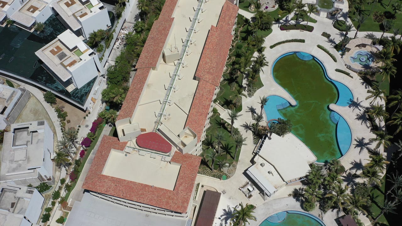 Overhead Los Cabos, Mexico view with a curved blue-edged pool, red tile roofs, palms, white buildings, nearby dark water, and no visible people.