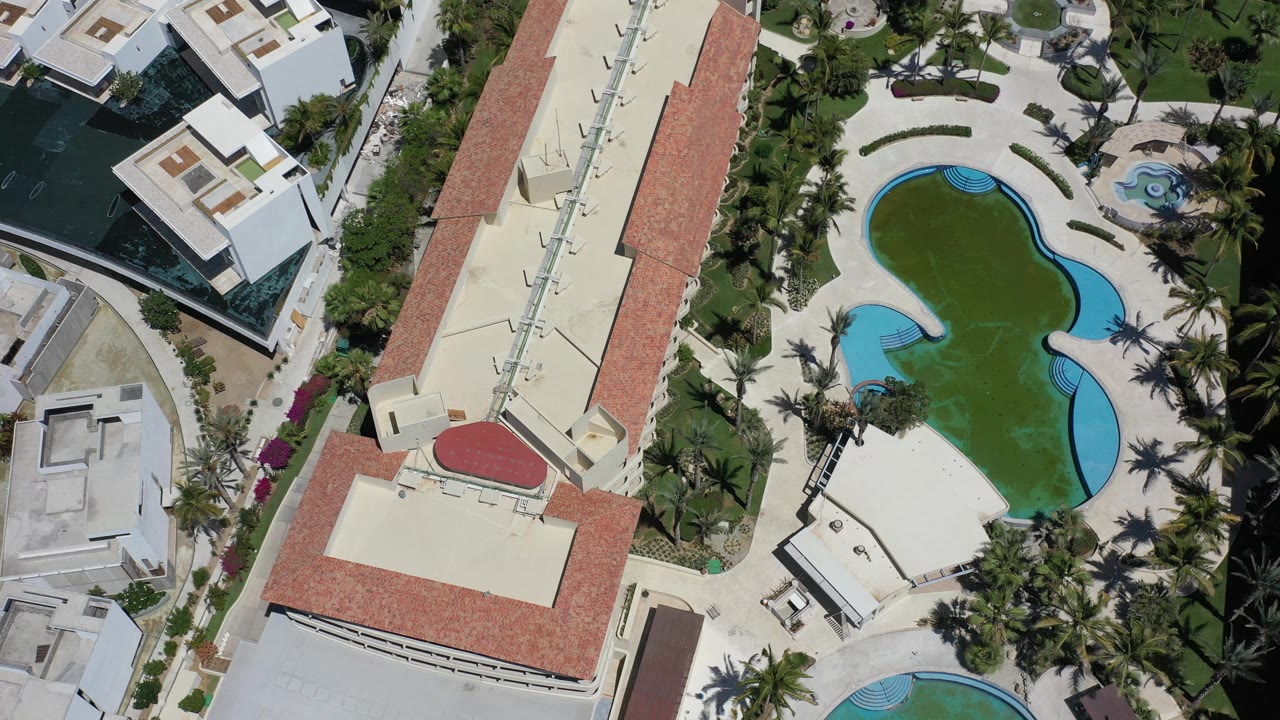 High overhead Los Cabos, Mexico frame centered on a long red-roofed building, a curved pool with green water, palms, pale paving, and no visible people.