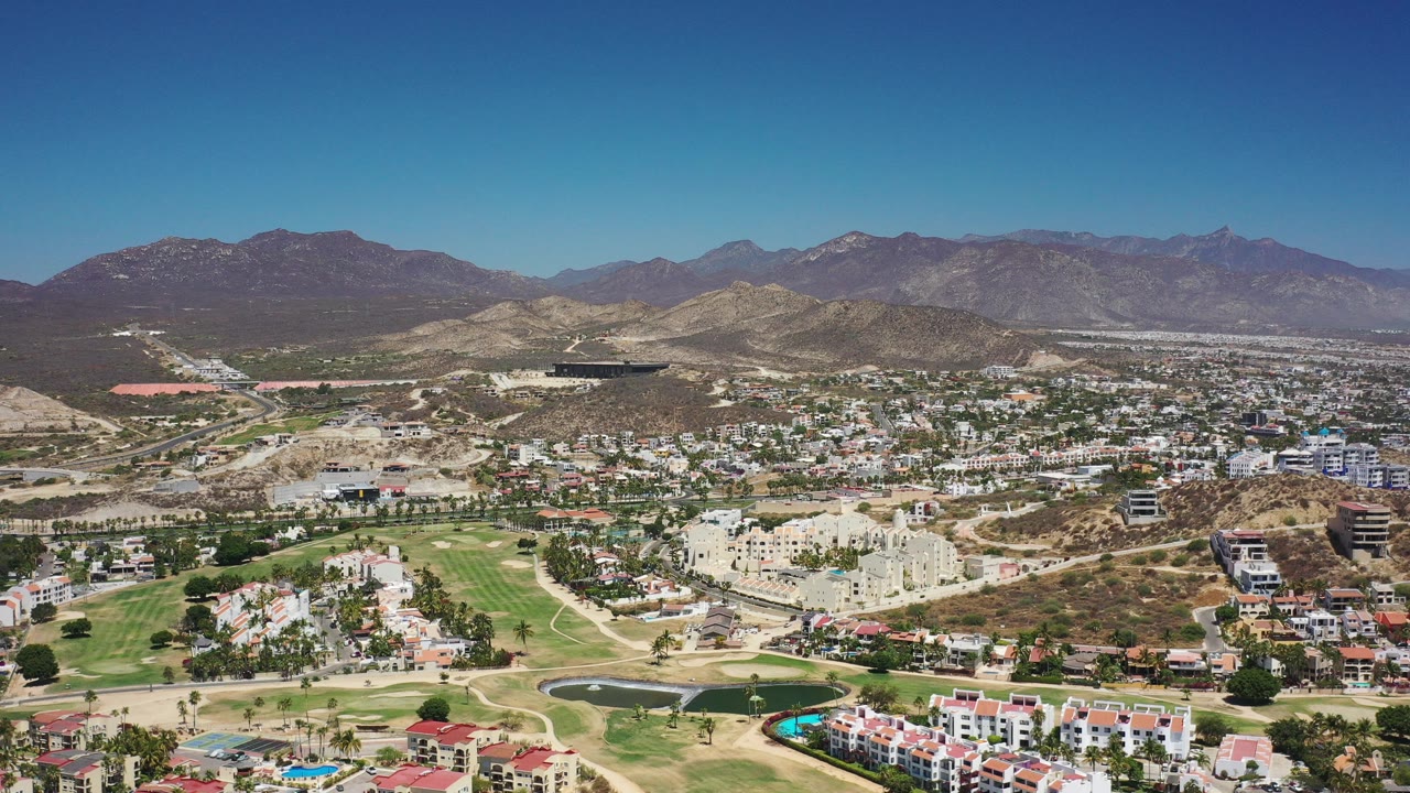 Opening aerial frame of Los Cabos, Mexico, showing a hillside town, green fairways, dry ridges, distant mountains, and no visible people.