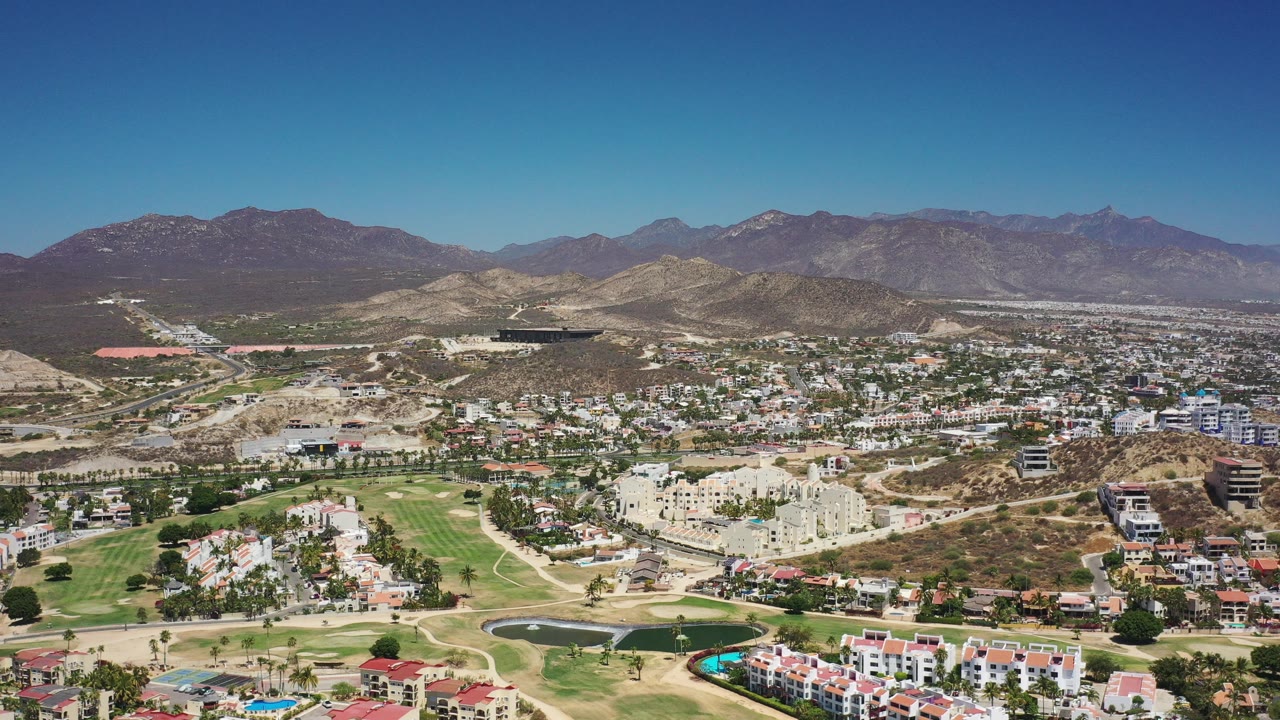 Aerial view of homes, green grounds, dry hills, and mountains under blue sky.