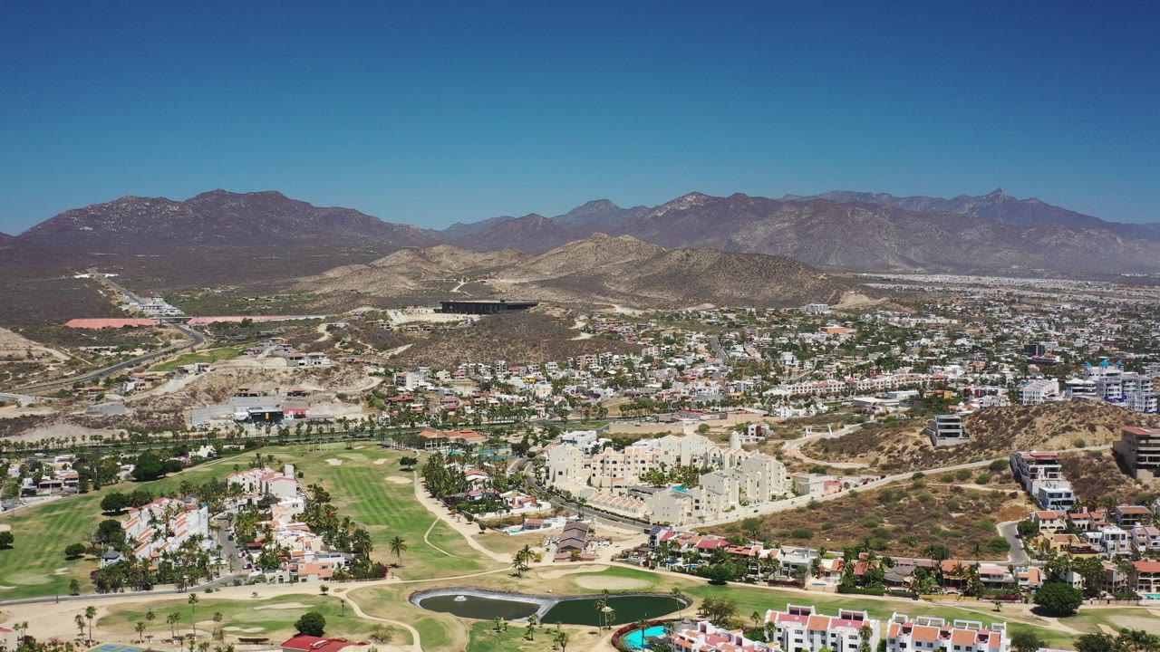Aerial view of a Los Cabos city area with golf greens, buildings, hills, and mountains.