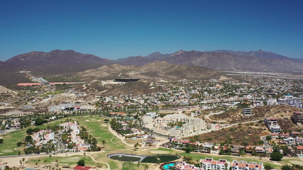 Los Cabos aerial view with fairways and a dark pond low in frame, residential buildings across the center, tan hills behind them, mountains on the horizon, and no visible people.