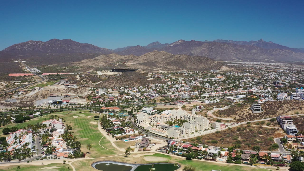 Sunlit aerial of Los Cabos buildings, roads, golf course, and mountain backdrop.