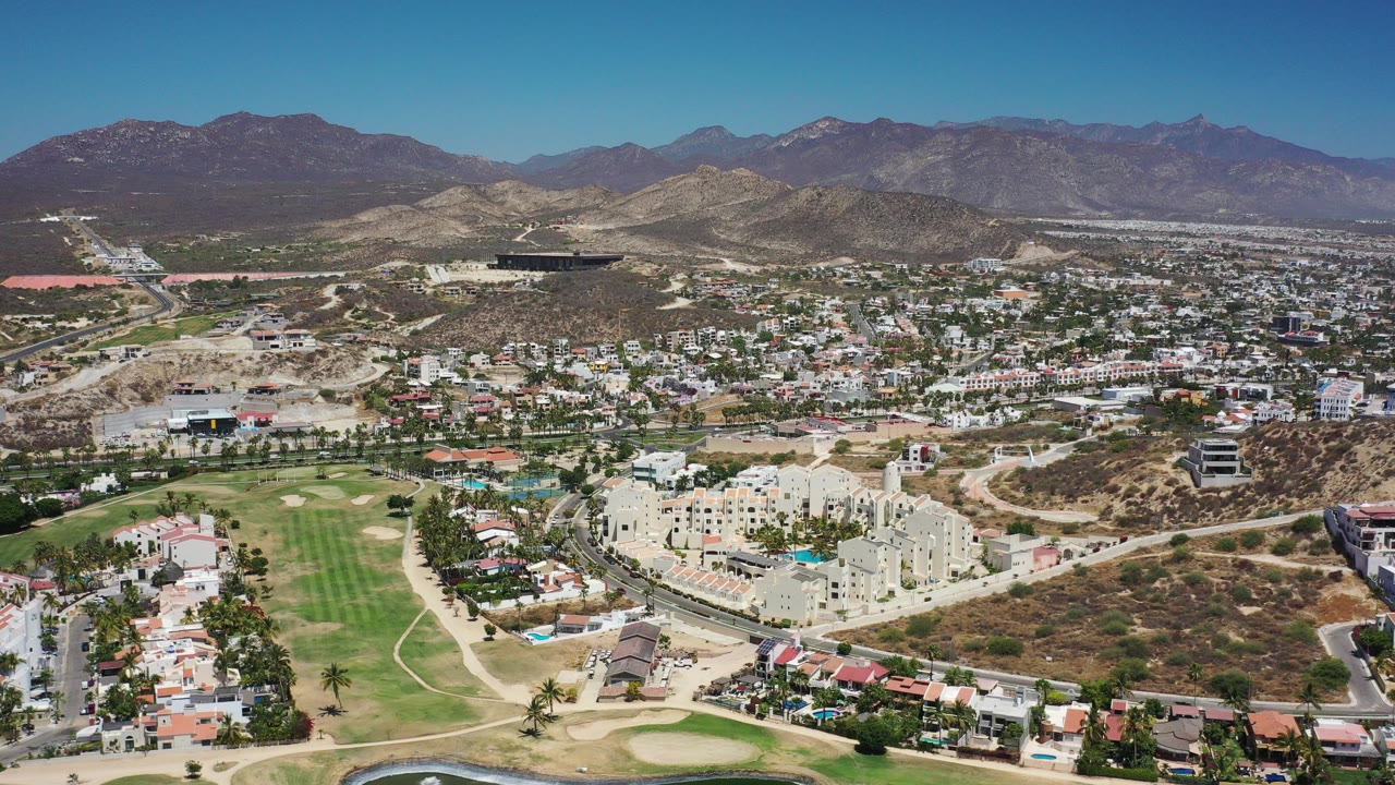 High aerial view over Los Cabos, Mexico, showing a green manicured course in the foreground, low-rise city buildings, dry hills, distant mountains, and no visible people.