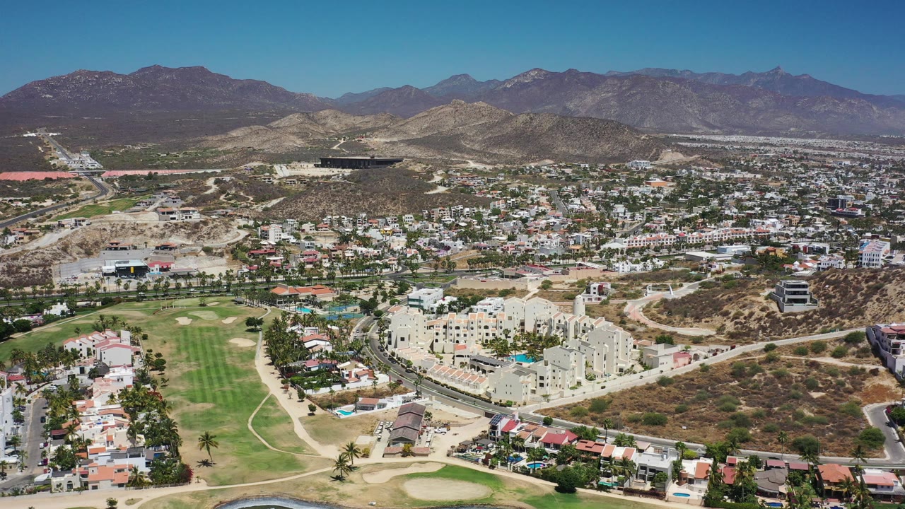 Wide aerial cityscape with course grass, buildings, hills, and mountain backdrop
