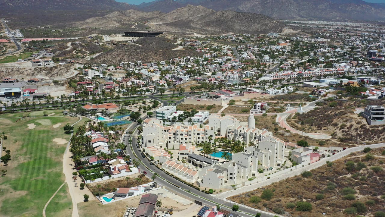 Opening aerial frame over Los Cabos, Mexico, showing cream resort-style buildings beside a curved road, green fairway, scattered homes, dry hills, and no visible people.