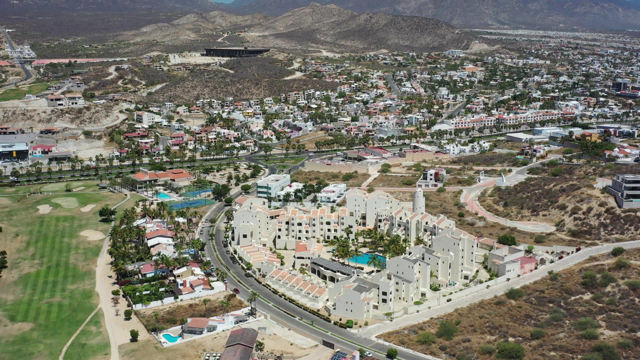 Drone view over pale buildings, road curves, fairway, and hillside homes.