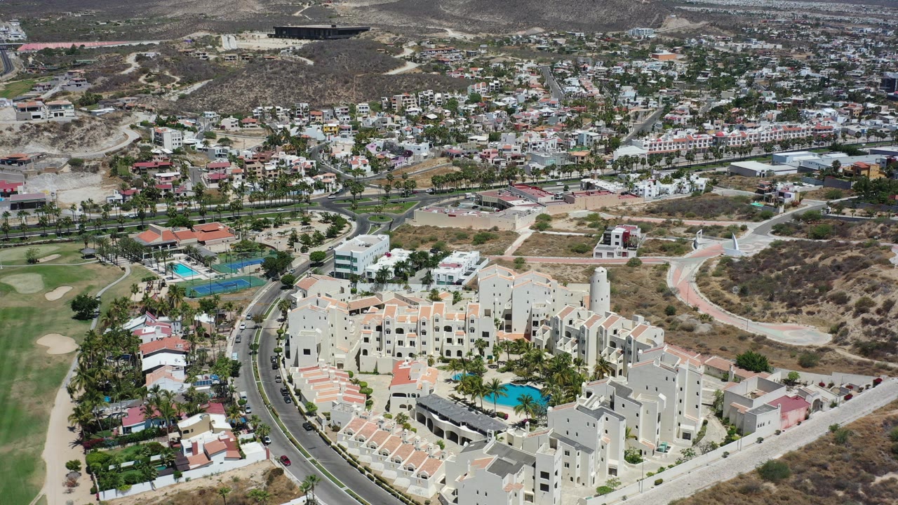 Aerial view over cream residential buildings, pools, roads, golf grass, and dry hills.