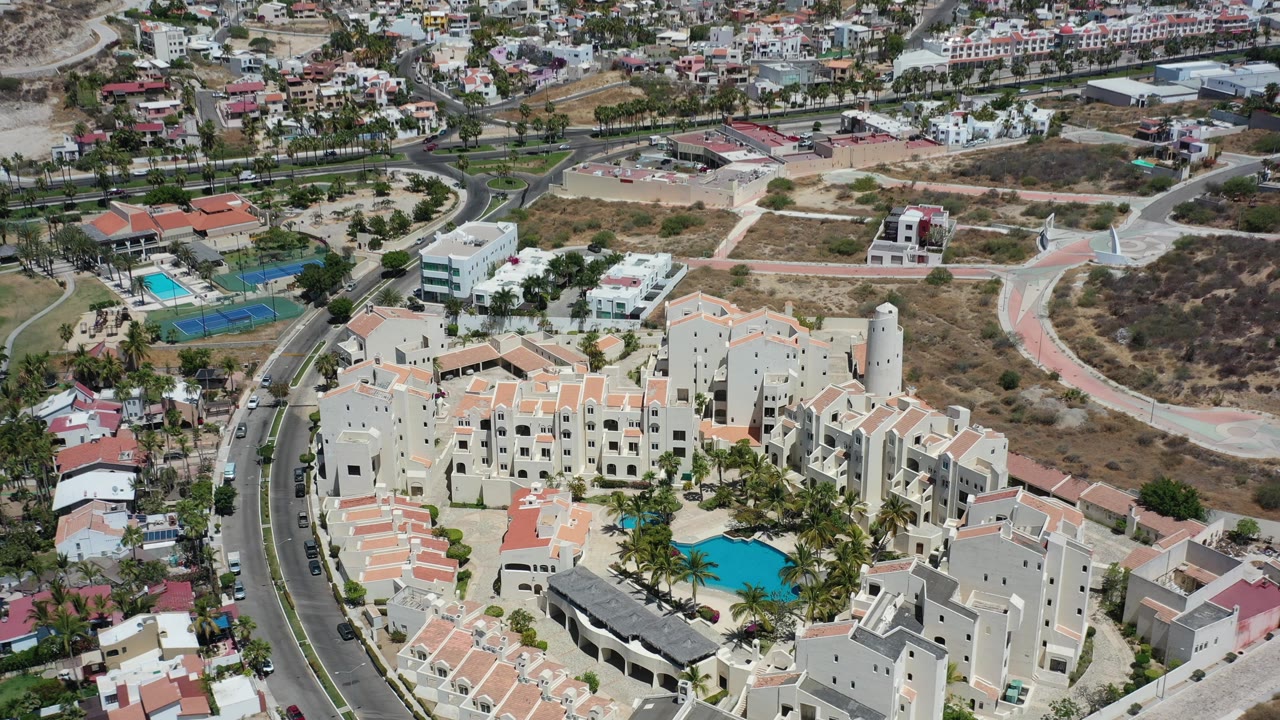Aerial frame of the Los Cabos resort from above, with the pool shifted near the lower right, pale hotel blocks in the center, a curved road at left, tennis courts beyond, and no visible people.