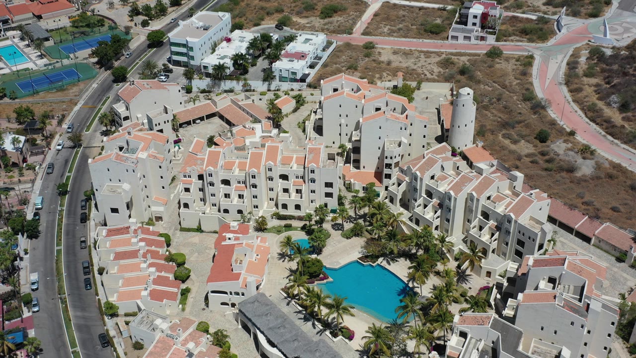 Aerial view of a resort complex with pool, palms, red tile roofs, roads, and dry terrain.