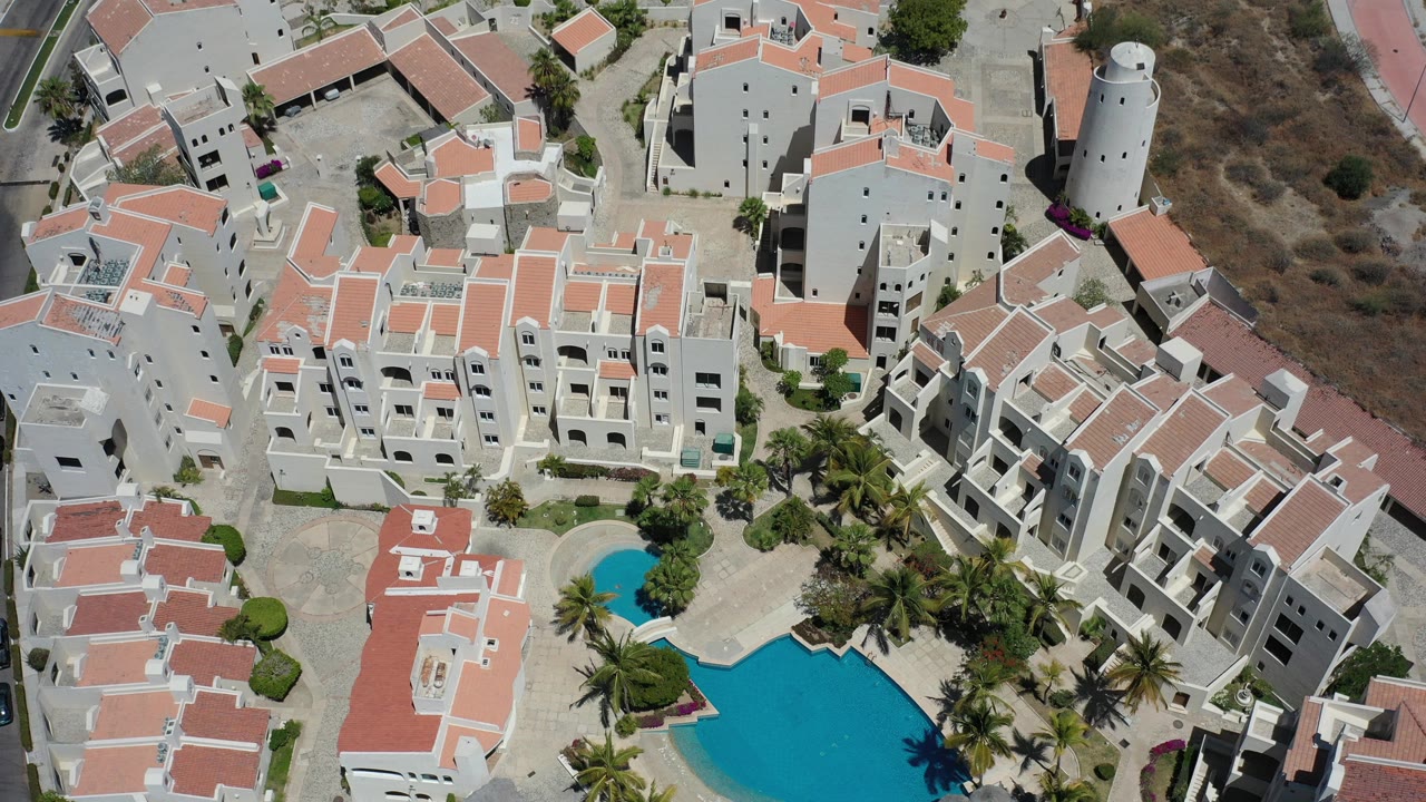 Aerial view of white condominium buildings, red roofs, palm trees, and a blue pool.