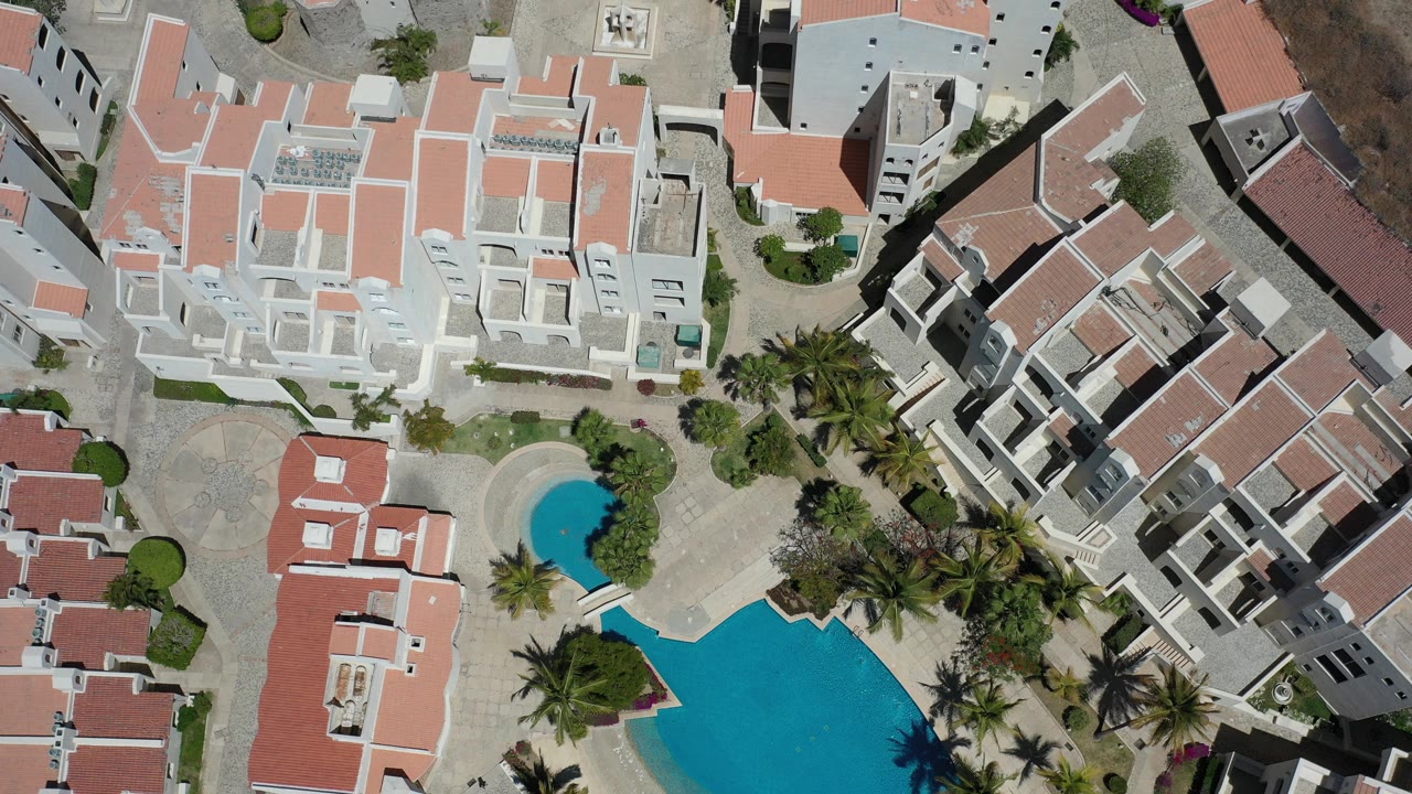 Overhead aerial frame in Los Cabos, Mexico showing white residential buildings with red roofs, two blue pools, palm trees, paved paths, and no visible people.