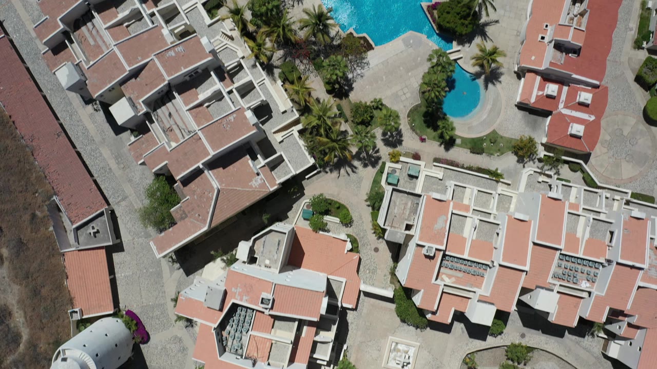 Overhead Los Cabos frame with pale building roofs, a large blue pool near the top edge, a smaller round pool, palms, paved courtyards, and no visible people.
