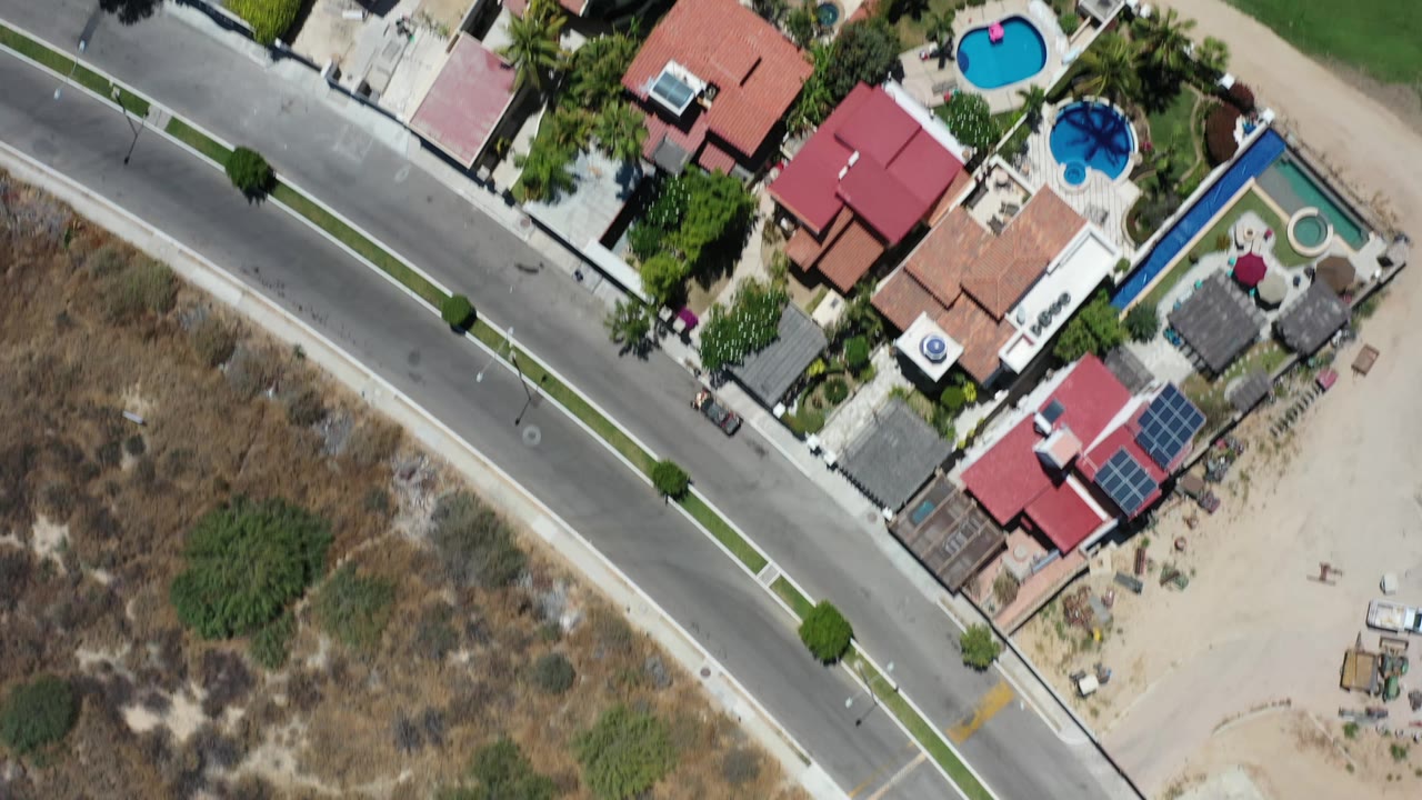 Overhead Los Cabos aerial view with tile-roof homes, multiple backyard pools, a straight road, scrubby dry ground, trees, and no visible people.