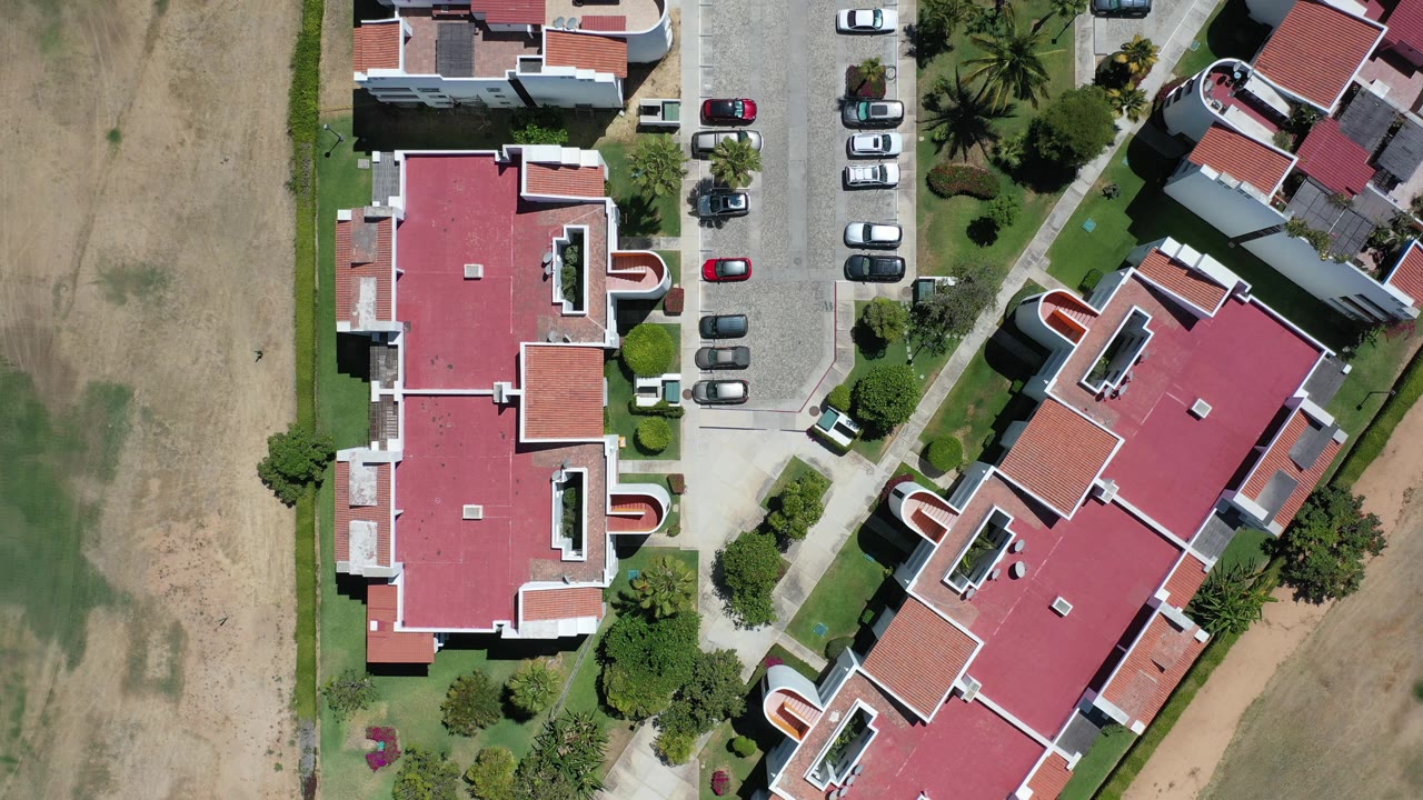 Top-down aerial view of red-roofed buildings beside dry open ground and parked cars.