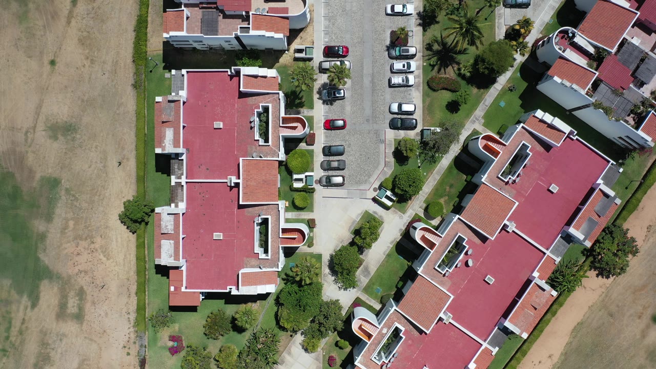 Overhead Los Cabos frame with the red-roofed buildings centered between a dry lot and parking spaces, with no visible people in the scene.