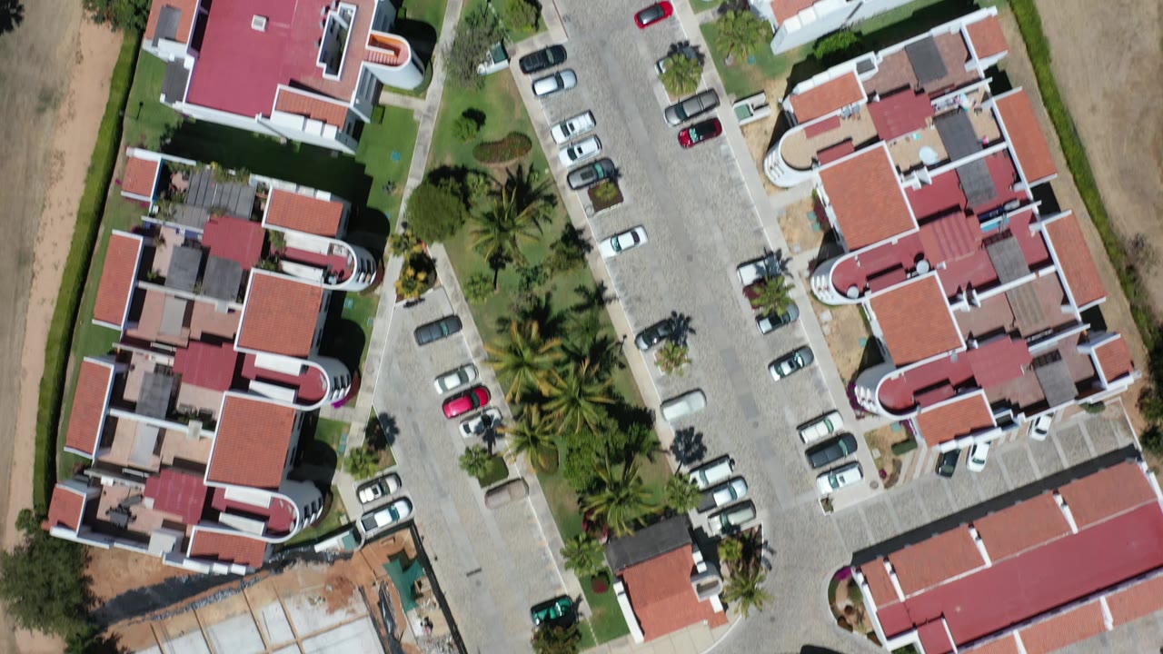 High overhead frame in Los Cabos, Mexico with the hotel roofs centered between a paved road, parked cars, palm landscaping, and tan desert ground, with no visible people.