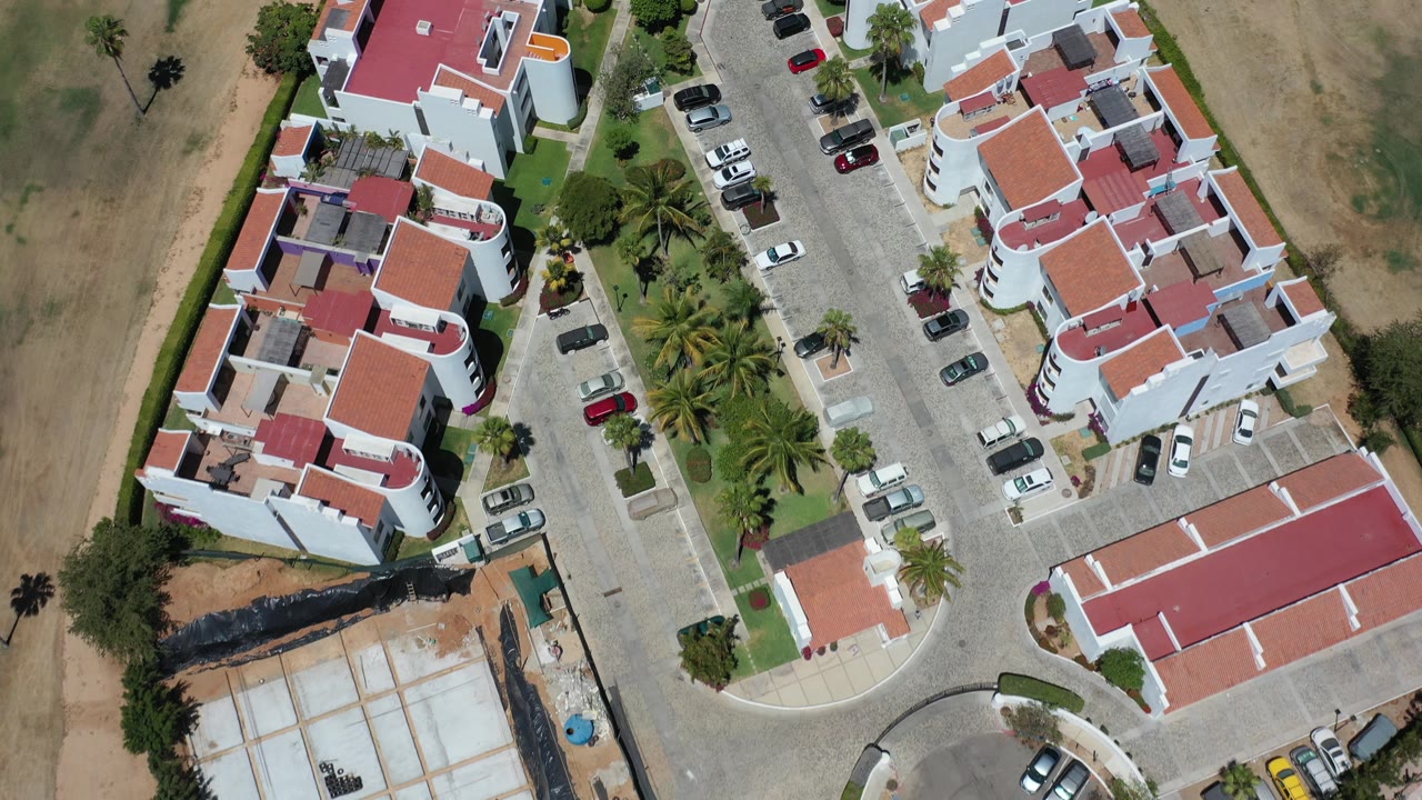 Overhead Los Cabos view of white residential buildings with red tile roofs, parked cars, palm trees, dry ground, and no visible people at the start of the clip.