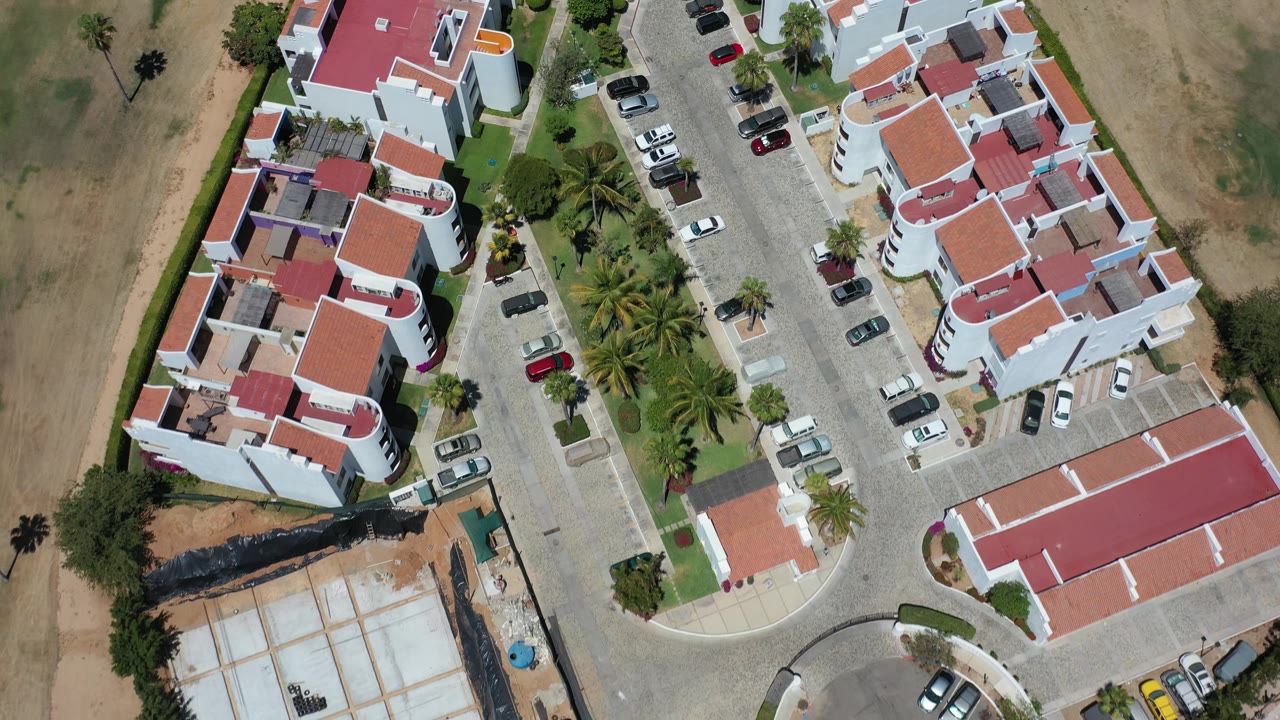 Los Cabos aerial frame looking down on red-roofed white buildings, a narrow internal road, parked vehicles, landscaped palms, and no visible people.