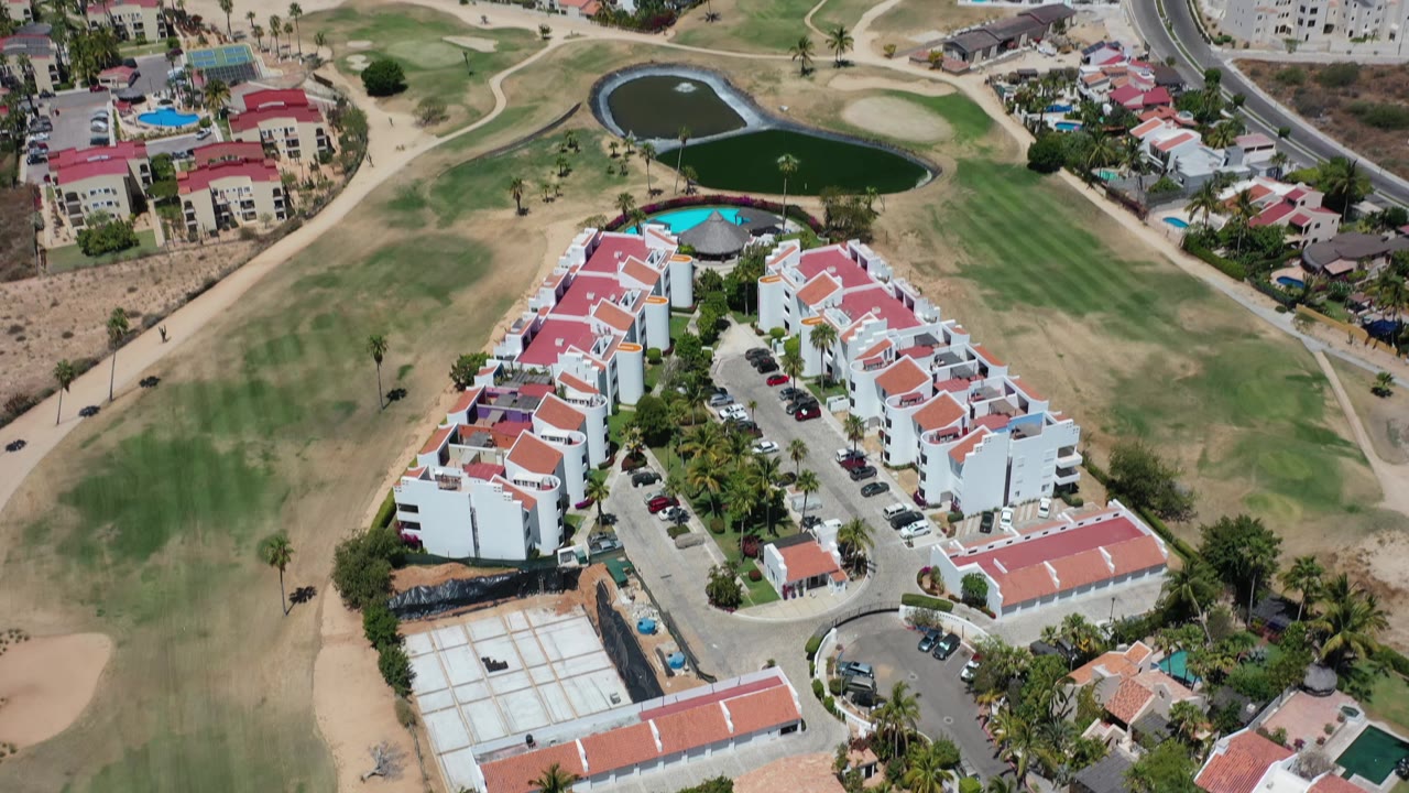 Aerial view in Los Cabos, Mexico, centered on red-roofed housing blocks with a parking lane, nearby golf greens, two dark ponds, and no visible people.
