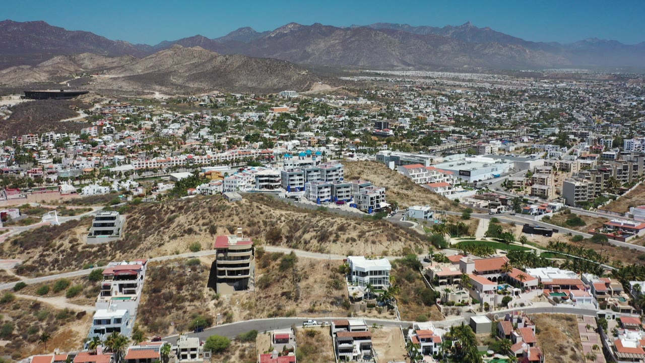 Aerial view over dry hills, houses, city blocks, and distant mountains.