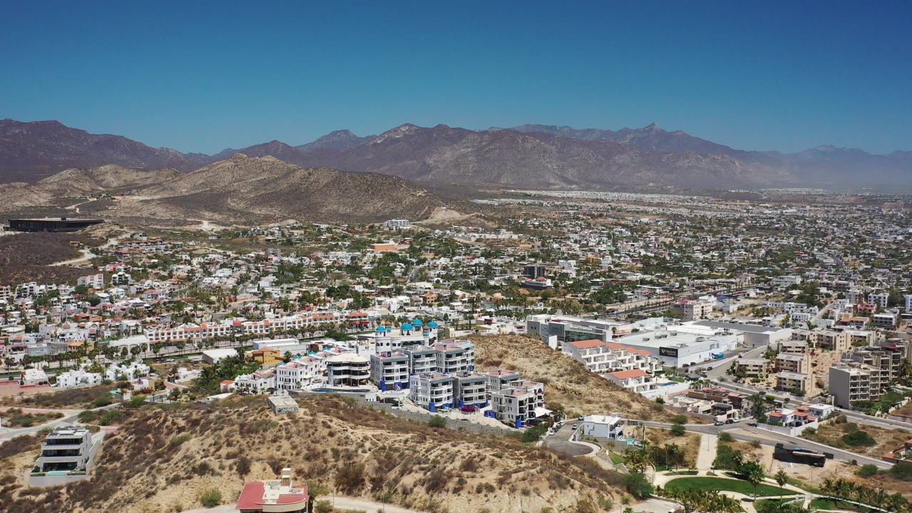 Opening frame of Los Cabos with scrubby foreground hills, low-rise buildings, distant mountains, blue sky, and no visible people.
