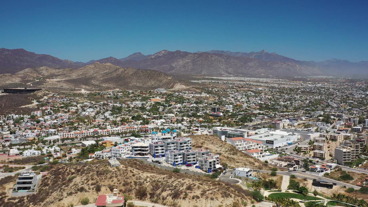 Los Cabos city blocks fill the midground beneath rugged mountains, with dry hillside terrain in front and no visible people.