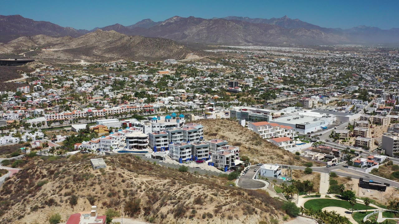 Aerial view over dry hills, city buildings, roads, and distant mountains.