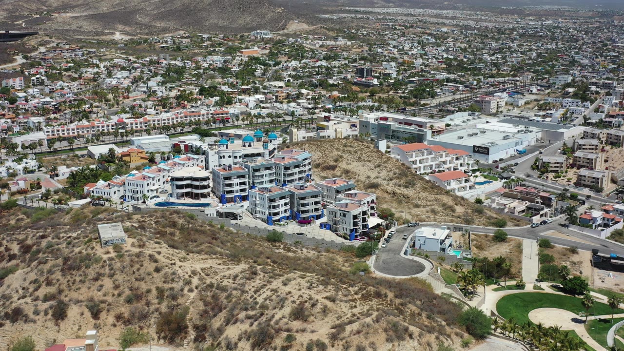 Early aerial frame over Los Cabos, Mexico, showing a curving road beside sandy hillside, white apartment-style buildings, city development, distant mountains, and no visible people.