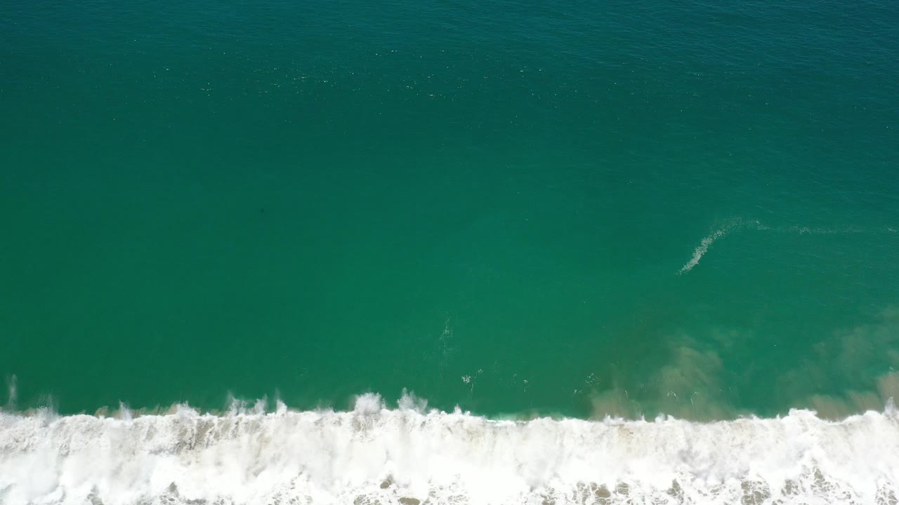 High overhead view of teal ocean water in Los Cabos, Mexico, with a white breaking wave line across the lower frame and no visible people.