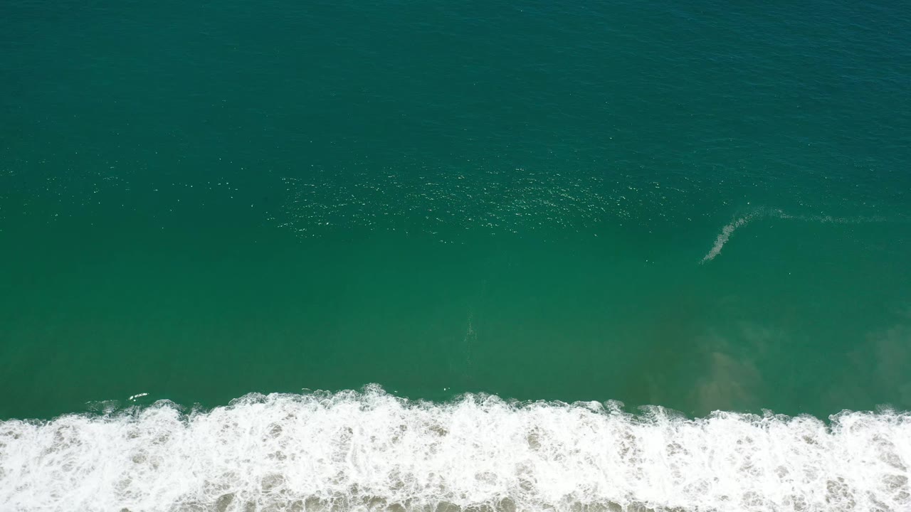 Aerial view of Los Cabos ocean water with bright white foam spreading horizontally across the lower third and no visible people.