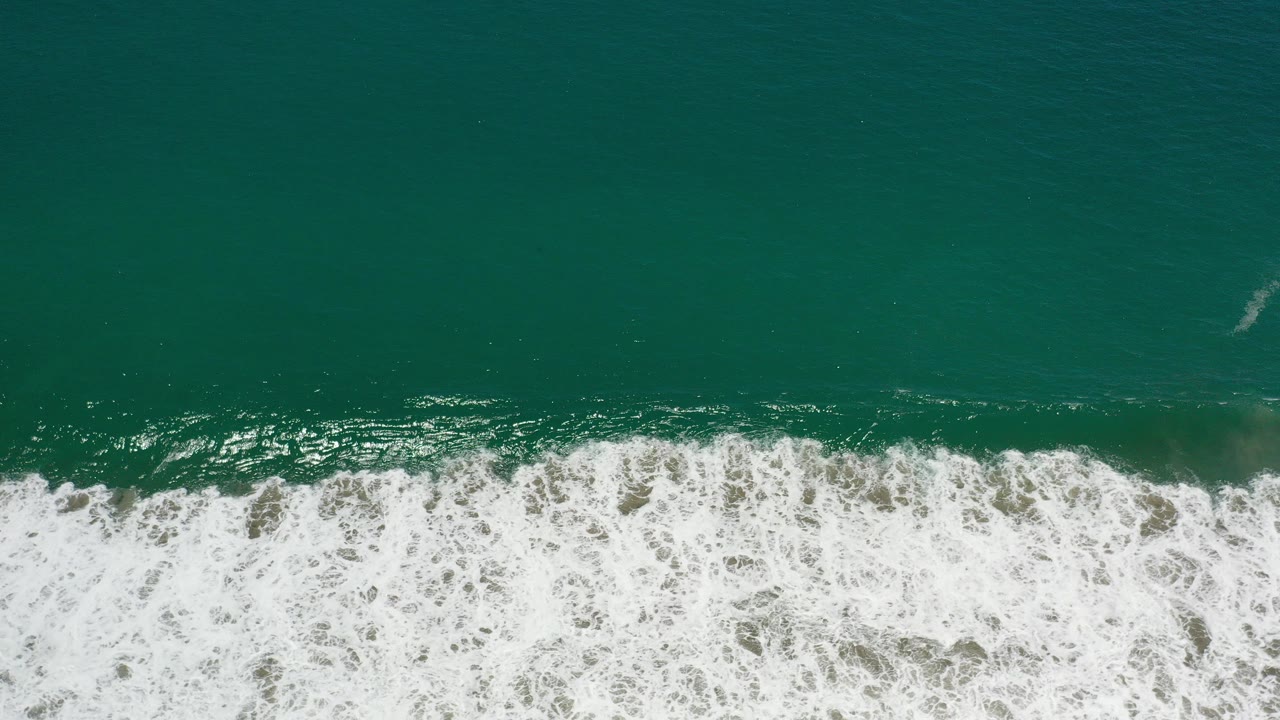 Top-down view of green water and a wide white surf band along a Los Cabos beach, with no visible people.