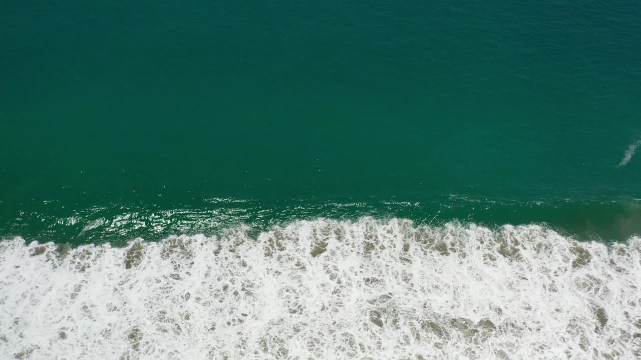 Aerial frame of Los Cabos green ocean water as white foam spreads across the lower shoreline, with no visible people.