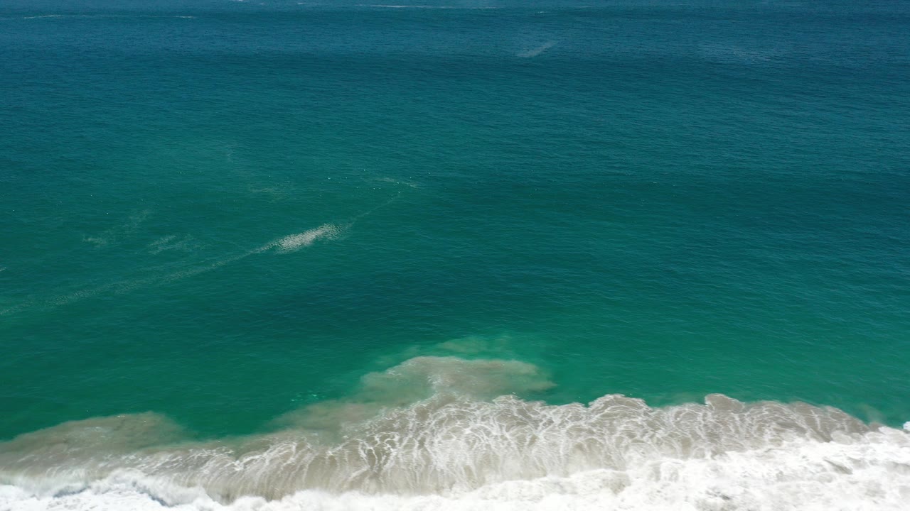 Aerial view of turquoise and deep blue ocean off Los Cabos, Mexico, with white surf foaming near the bottom edge and no visible people.