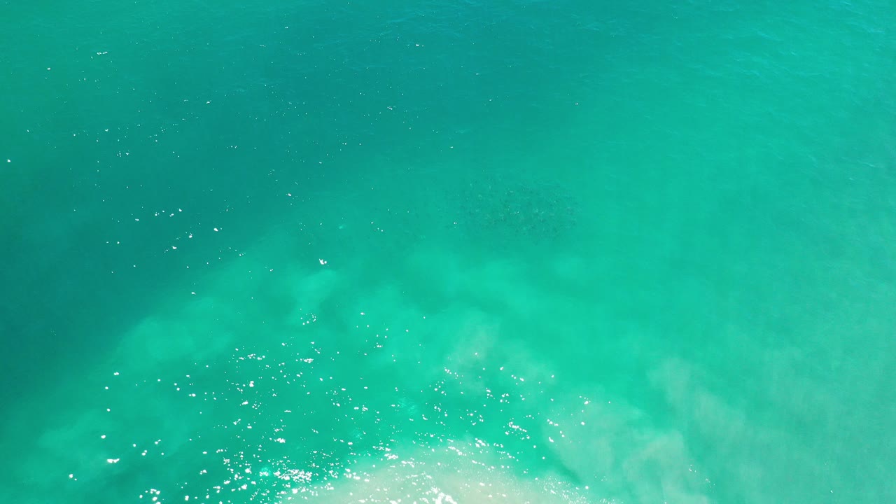 Overhead view of turquoise and green ocean water in Los Cabos, Mexico, with pale wave wash entering near the lower edge and no visible people.