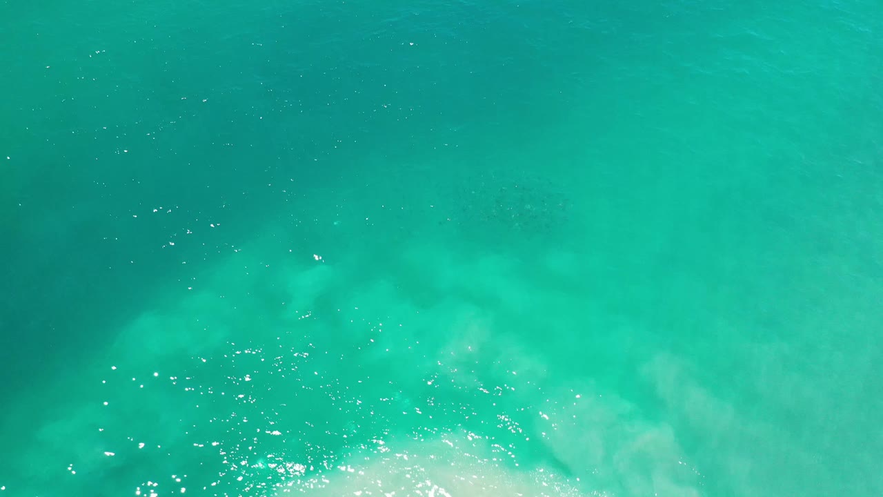 Blue-green ocean surface in Los Cabos, Mexico, seen from high above as white turbulence spreads across the lower part of the frame with no visible people.