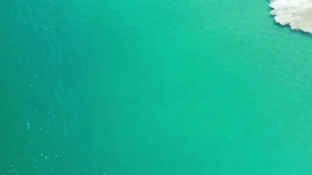 Overhead view of clear green ocean in Los Cabos, Mexico, with a small patch of white surf at the upper right and no visible people.