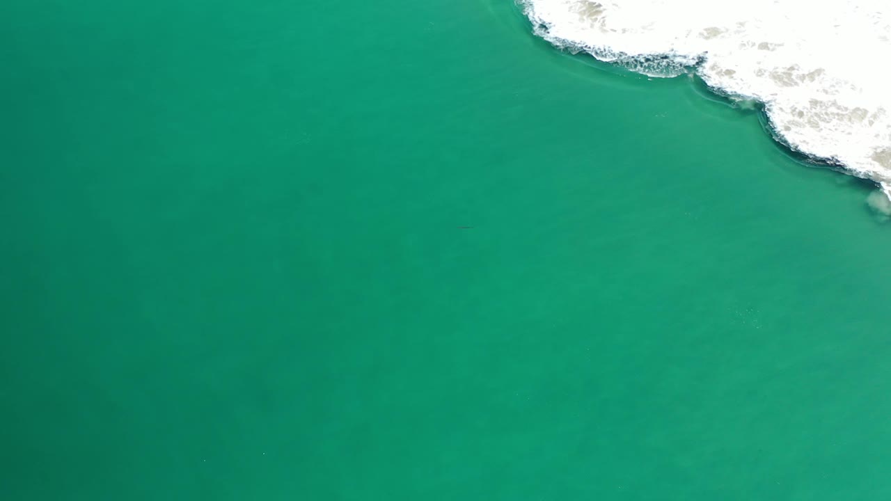 Aerial view of blue-green Los Cabos water with white wave foam crossing the top edge and faint darker water texture below, with no visible people.