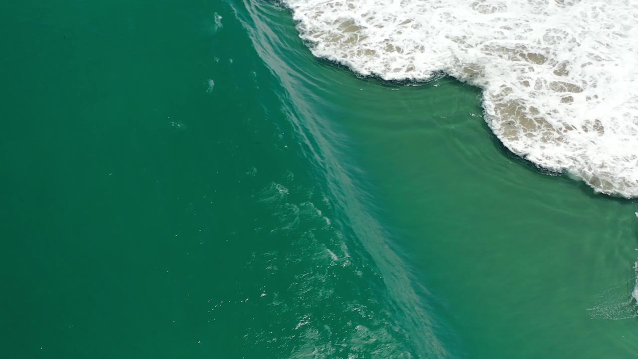 Aerial Los Cabos ocean frame with a white foam line spreading along the top edge over blue-green water and no visible people.