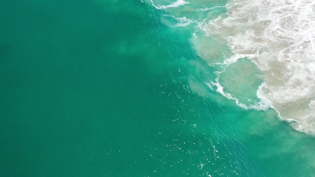 Overhead view of turquoise sea in Los Cabos, Mexico, with white surf spreading over pale sandy shallows at the upper right and no visible people.