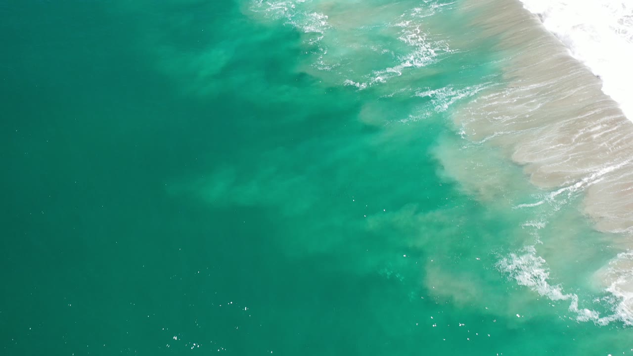Overhead view of emerald green water and pale surf along the Los Cabos, Mexico shoreline at the start of the clip, with no visible people.