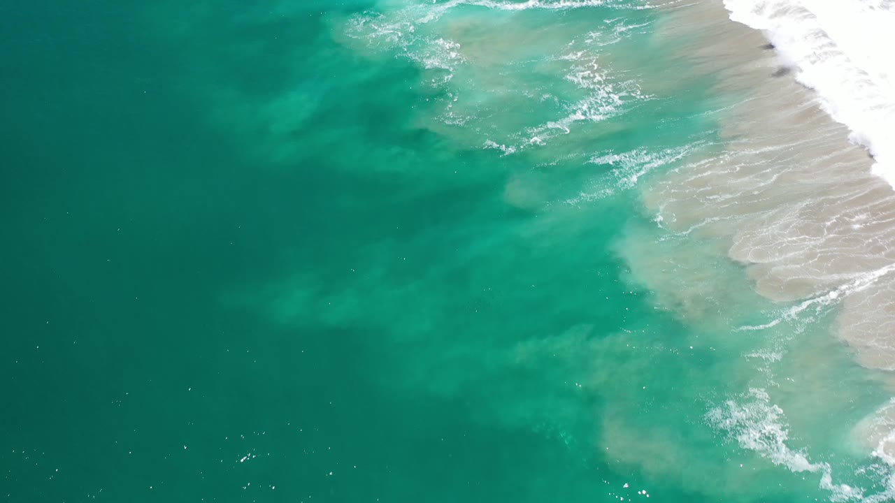 Turquoise ocean seen from above with surf breaking along the upper right shoreline.