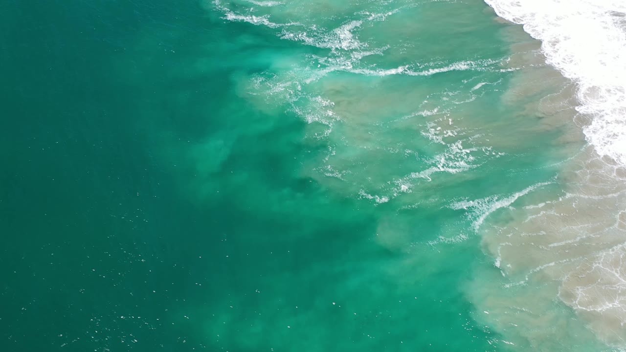 Aerial Los Cabos water view with darker ocean on the left, pale submerged sand on the right, and scattered foam lines across the shallows, no visible people.